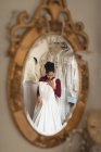 Reflexão da noiva tentando vestido de noiva de cabide de roupas no espelho na boutique — Fotografia de Stock