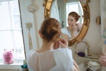 Young bride in wedding dress wearing earrings at boutique and looking in mirror on wall — Stock Photo