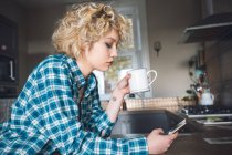 Женщина пьет кофе, используя мобильный телефон на кухне дома — стоковое фото