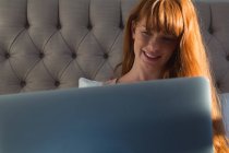 Mujer sonriente con el pelo rojo usando el ordenador portátil en el dormitorio en casa - foto de stock