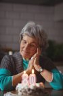 Задумчивая пожилая женщина с тортом на день рождения дома — стоковое фото
