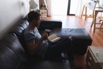 Hombre tomando café mientras lee un libro en la sala de estar en casa - foto de stock