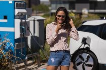 Femme parlant sur un téléphone portable tout en rechargeant une voiture électrique à la station de charge — Photo de stock