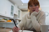 Thoughtful woman writing on diary at home in kitchen with hand on cheek — Stock Photo
