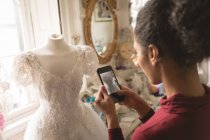 Femme métissée prenant des photos de robe de mariée sur téléphone portable en boutique — Photo de stock