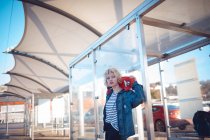 Mujer reflexiva sosteniendo monopatín en la parada de autobús - foto de stock