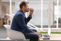 Бизнесмен пьет кофе во время использования ноутбука в офисе — стоковое фото