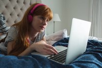 Frau benutzt Laptop mit Kopfhörer im heimischen Schlafzimmer — Stockfoto