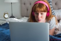 Femme utilisant un ordinateur portable avec écouteurs dans la chambre à coucher à la maison — Photo de stock