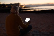 Старший мужчина использует цифровой планшет на пляже в сумерках — стоковое фото