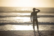 Vista trasera del surfista masculino de pie en traje de baño y arnés de cintura en la playa - foto de stock