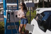 Mujer hablando por teléfono móvil mientras carga coche eléctrico en la estación de carga - foto de stock