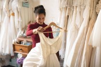 Mujer sonriente sosteniendo vestido de novia en percha de ropa en boutique - foto de stock