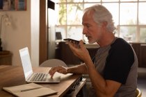 Homem sênior falando no telefone celular enquanto usa laptop em casa — Fotografia de Stock