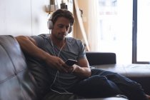 Homme utilisant un téléphone portable avec écouteurs dans le salon à la maison — Photo de stock