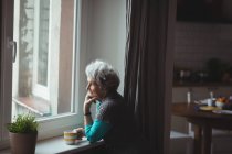 Старша жінка дивиться крізь вікно, маючи каву вдома — стокове фото