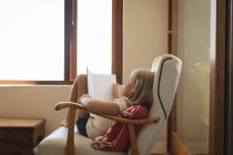 Дівчина навчається у вітальні вдома, сидячи в кріслі з книгою — стокове фото