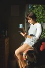 Женщина пьет кофе, используя мобильный телефон в гостиной дома — стоковое фото