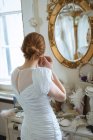 Young bride in wedding dress wearing earrings at boutique and looking in mirror on wall — Stock Photo