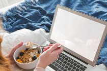 Frau mit roten Haaren benutzt Laptop im Schlafzimmer mit Schüssel-Frühstück — Stockfoto