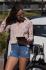 Женщина с цифровым планшетом во время зарядки электромобиля на зарядной станции — стоковое фото