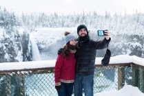 Casal tirando selfie com telefone celular perto de cachoeira durante o inverno — Fotografia de Stock