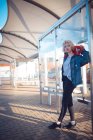 Joven mujer sosteniendo monopatín en parada de autobús - foto de stock