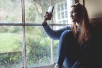 Жінка бере селфі з мобільним телефоном біля вікна вдома — стокове фото