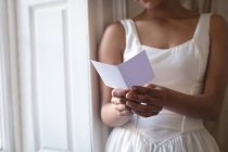 Vue partielle de la mariée en robe de mariée lecture carte de vœux à la fenêtre — Photo de stock