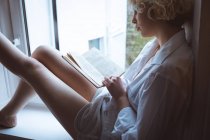 Femme lisant un livre près de la fenêtre à la maison — Photo de stock