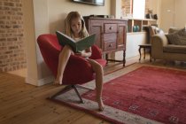 Chica estudiando en casa y sentada en sillón con libro - foto de stock