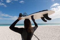 Rear view of male surfer carrying surfboard in the beach — Stock Photo