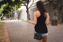 Вид ззаду жінки за допомогою мобільного телефону під час ходьби на тротуарі — стокове фото