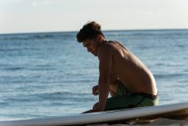 Молодий чоловічий серфер сидить з дошкою для серфінгу на пляжі — стокове фото