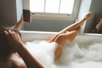 Woman taking selfie with mobile phone while taking bubble bath in bathroom — Stock Photo