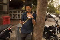 Улыбающийся бизнесмен, пользующийся мобильным телефоном во время еды — стоковое фото