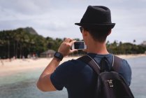 Вид сзади человека, щёлкающего фото с мобильного телефона рядом с пляжем — стоковое фото