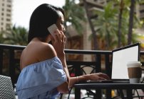 Жінка розмовляє на мобільному телефоні, використовуючи ноутбук у кафе — стокове фото
