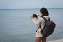 Vue arrière de la femme en cliquant sur la photo de la mer avec appareil photo numérique à la plage — Photo de stock