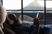 Старший мужчина лежит на диване, слушая музыку на смартфоне дома — стоковое фото