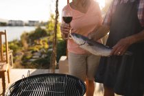 Sección media de la pareja mayor sosteniendo pescado y copa de vino en el patio trasero - foto de stock