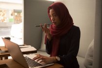 Femme d'affaires dans le hijab en utilisant un ordinateur portable tout en parlant sur un téléphone mobile à la cafétéria de bureau — Photo de stock