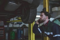Мужчина-механик разговаривает по мобильному телефону во время осмотра автомобиля в ремонтном гараже — стоковое фото