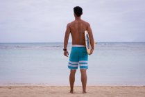 Вид ззаду чоловіка, що стоїть з дошкою для серфінгу на пляжі — стокове фото