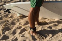 Surfista di sesso maschile in piedi con guinzaglio tavola da surf sulla gamba in spiaggia — Foto stock