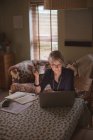 Femme mature utilisant un ordinateur portable tout en prenant un café dans le salon à la maison — Photo de stock