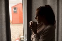 Mujer pensativa tomando café mientras mira a través de la ventana en casa - foto de stock