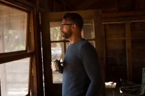 Man in log cabin with coffee mug looking through window — Stock Photo