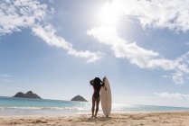Donna in piedi con tavola da surf in spiaggia in una giornata di sole — Foto stock