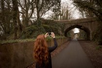 Вид ззаду жінки, що клацає фотографією з мобільним телефоном на сільській дорозі — стокове фото